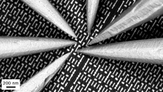 SEM close-up of six probe tips converging radially on a patterned chip surface, 200 nm scale bar.