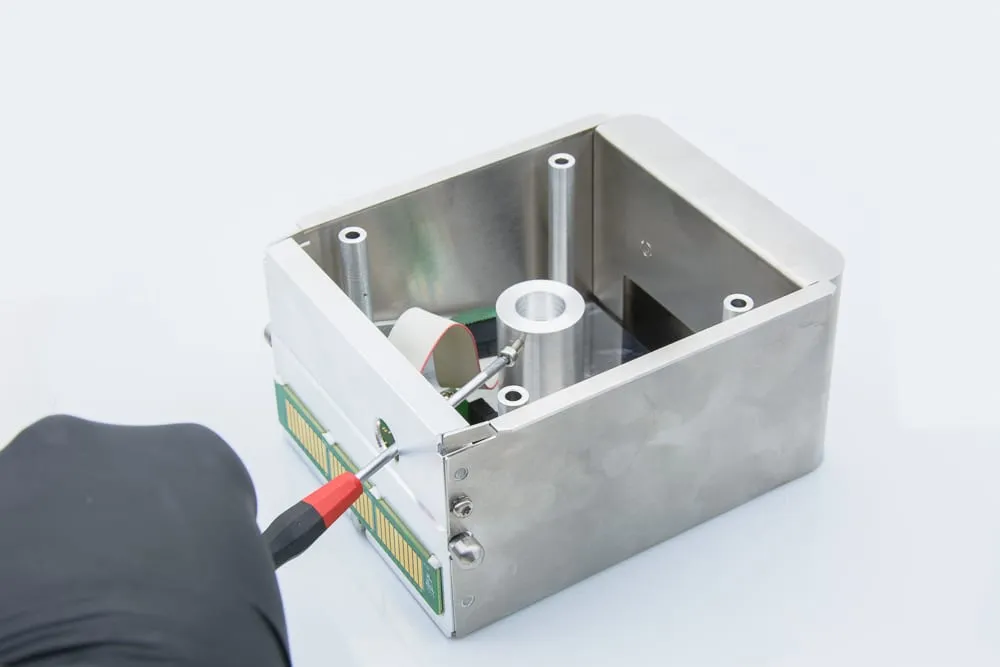 The Prober Shuttle chassis open and empty, gloved hand and screwdriver positioning it for assembly, with mounting posts and a gold flex-circuit connector visible inside.