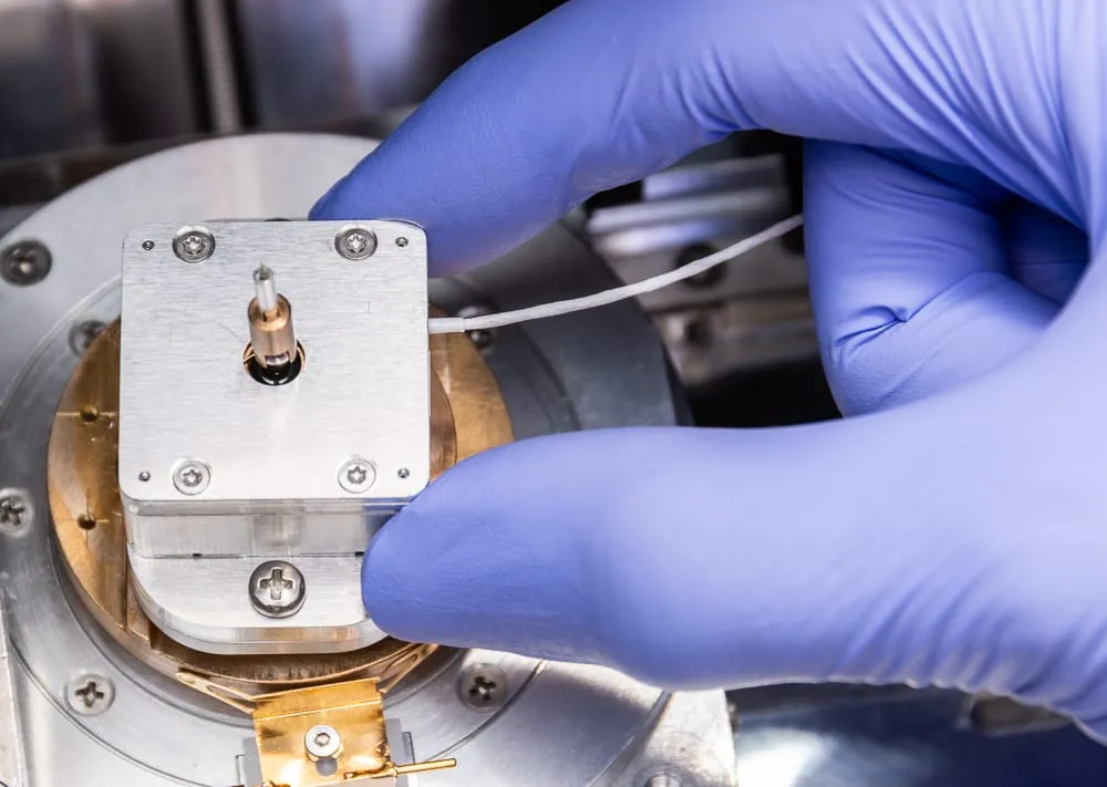 A gloved hand adjusting the nanolathe's collet and fine pin, showing the unit is small enough to hold and align by hand.